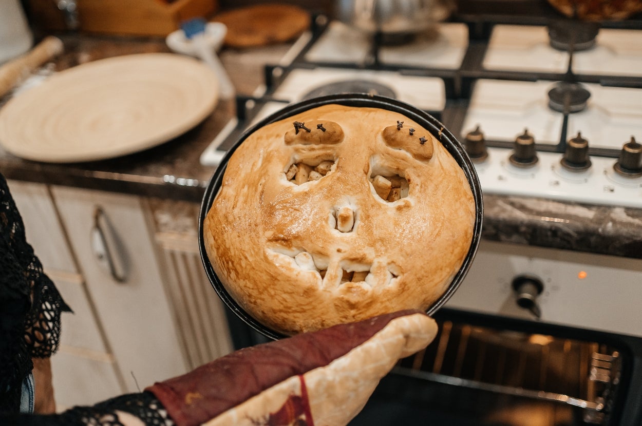 A pie with a spooky face carved on top is held on a gloved hand in a kitchen setting