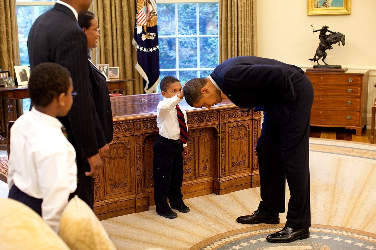 A young boy touches the head of a man in a suit who is bending over toward him in an office setting. Three other adults look on