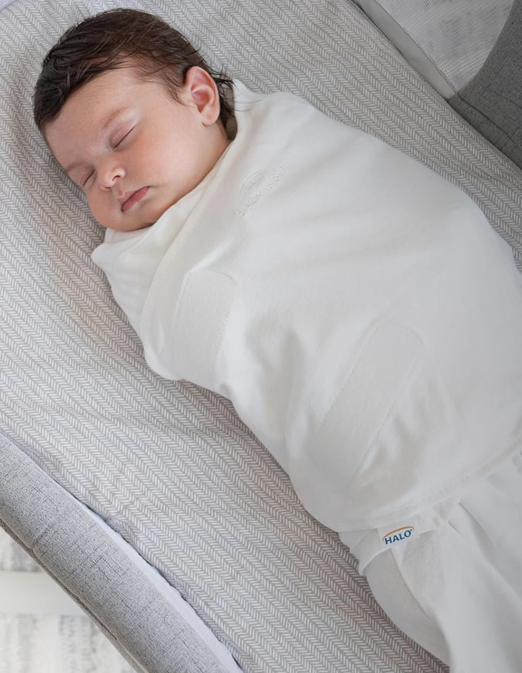 Sleeping baby wrapped in a soft white swaddle on a patterned surface, promoting baby sleep products