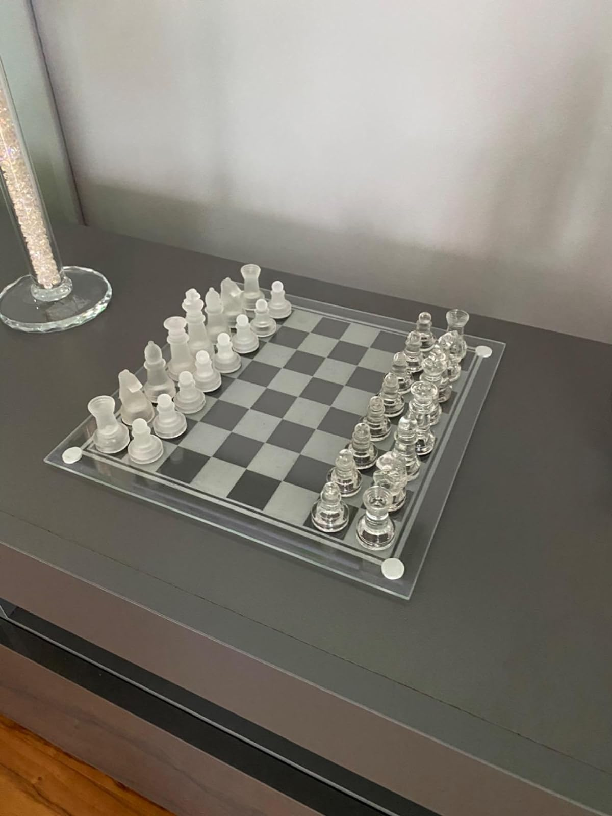 A clear glass chess set arranged for the start of a game on a dark table