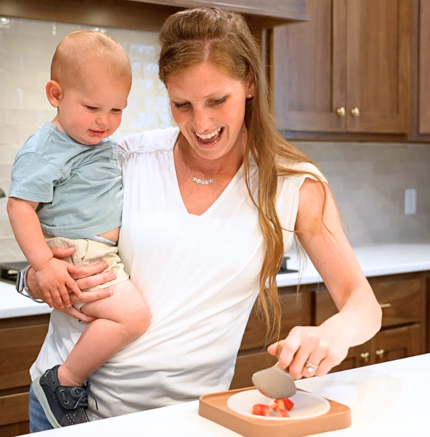 Woman holding a child uses a utensil to prepare sliced food on a plate in a kitchen