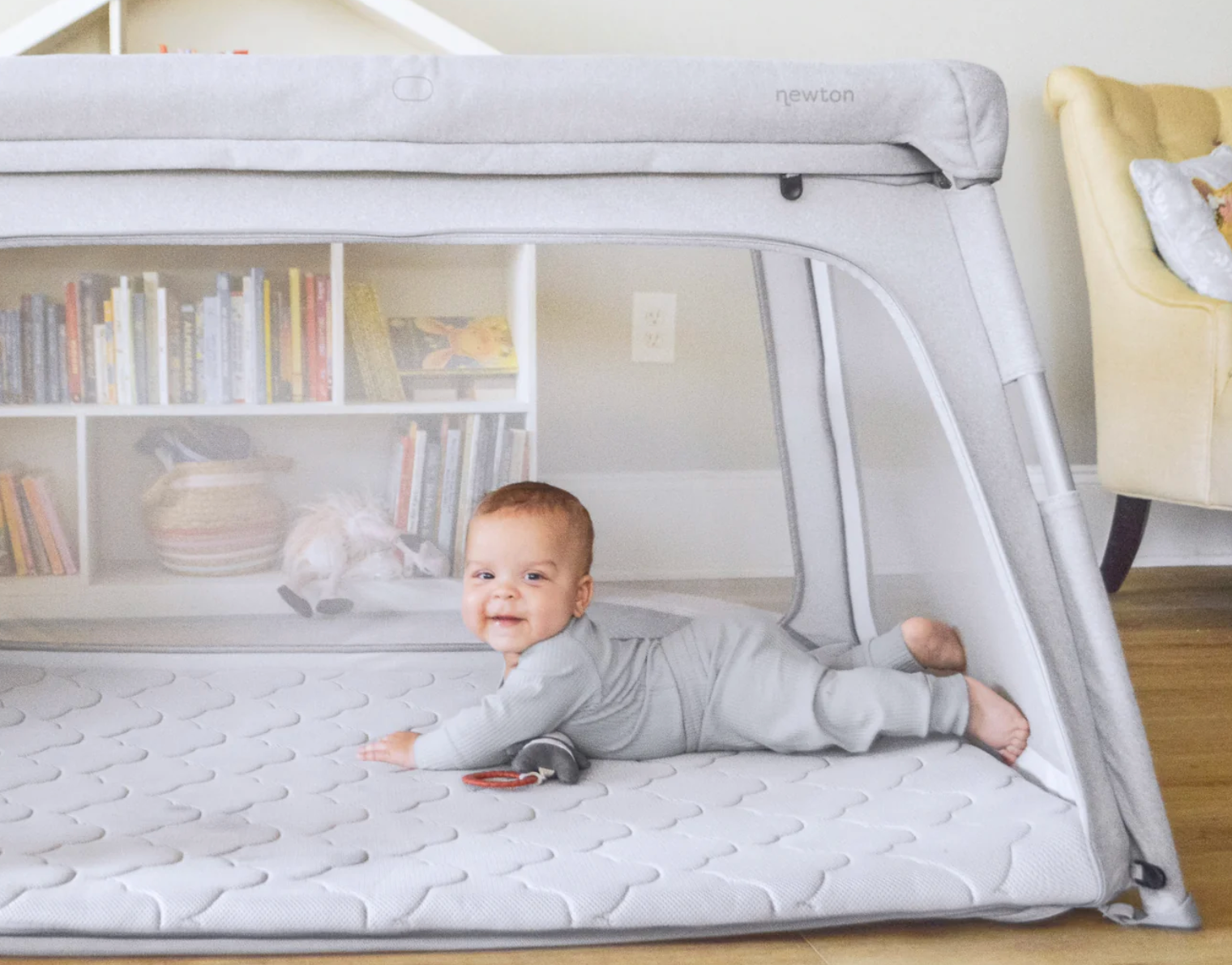 Baby smiling on a padded mat inside a mesh playpen, with a bookshelf and toys in the background
