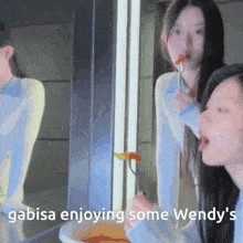 two girls are eating food in front of a mirror with the words " gabisa enjoying some wendy 's " below them