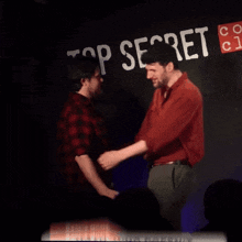 two men shake hands in front of a top secret sign