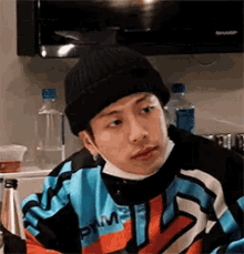 a young man wearing a black beanie and a blue jacket is sitting in a kitchen .