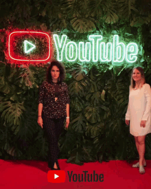 two women standing in front of a youtube neon sign