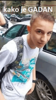 a young man with a backpack is standing in front of a car with the words kako je gaban above him .