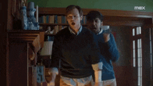two men are dancing in a living room with a max logo in the corner .