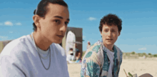 two young men are sitting on a beach and one is wearing a pearl necklace