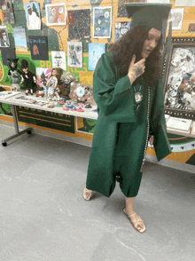 a woman in a green graduation cap and gown stands in front of a table with artwork on it