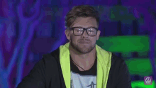 a man wearing glasses and a yellow jacket is sitting in front of a neon sign that says mega