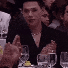 a man in a black suit is sitting at a table with glasses of water and clapping his hands .