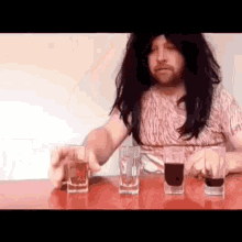 a man in a wig is sitting at a table with four shot glasses of alcohol .