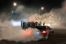 a group of people are standing in front of a car that has smoke coming out of its tires