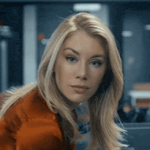 a close up of a woman 's face with long blonde hair wearing an orange jacket .
