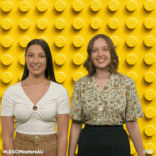 two women standing in front of a yellow wall with lego bricks