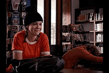 a girl wearing an orange shirt and a black beanie is sitting in a library