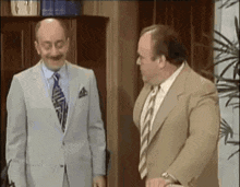 two men in suits and ties are standing next to each other in an office and talking .