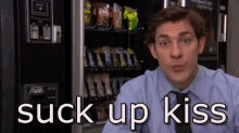 a man in a suit and tie is sitting in front of a vending machine that says " suck up kiss " .