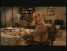 a bald man is sitting at a table in a living room with a bookshelf in the background .