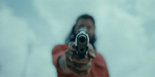 a man pointing a gun at the camera with a blue sky in the background