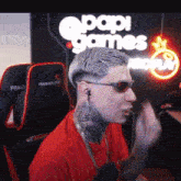a man wearing sunglasses and a red shirt sitting in front of a sign that says papi games