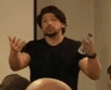 a man in a black shirt is talking to a group of people while holding a cell phone .