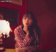 a woman sits at a table with her hand on her chin in front of a lamp with honeycam.org written on the bottom