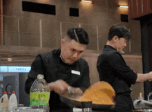 two men cooking in front of a pantry with a bottle of water in front of them