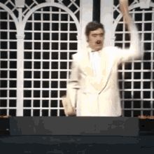 a man in a white suit and tie is standing in front of a fence and raising his arms in the air .
