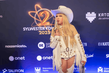 a woman in a cowboy outfit is standing in front of a sign that says sylwester zdv 2025