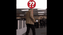a man in a tan jacket is standing in a room with a red question mark on his head
