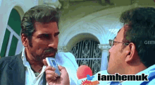 a man holding a cell phone talking to another man with the words iamhemuk on the bottom right