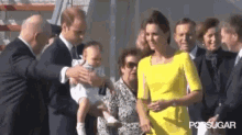 a man in a suit is holding a baby in his arms while a woman in a yellow dress looks on .