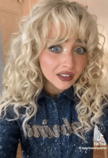 a woman with blonde curly hair and blue eyes is wearing a denim shirt and making a funny face .
