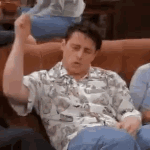 a man in a shirt is sitting on a couch with his hand up .