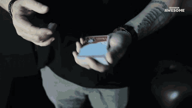a man with a tattoo on his arm is holding a pack of playing cards in his hand with the awesome logo on the bottom right