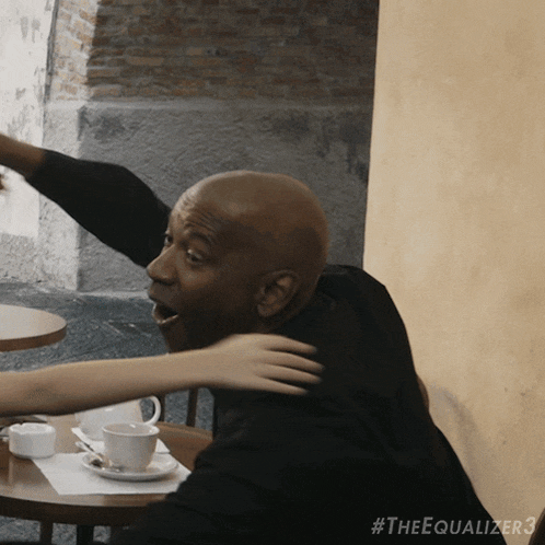 a man sitting at a table with a cup of coffee and a woman hugging him with #theequalizer3 on the bottom