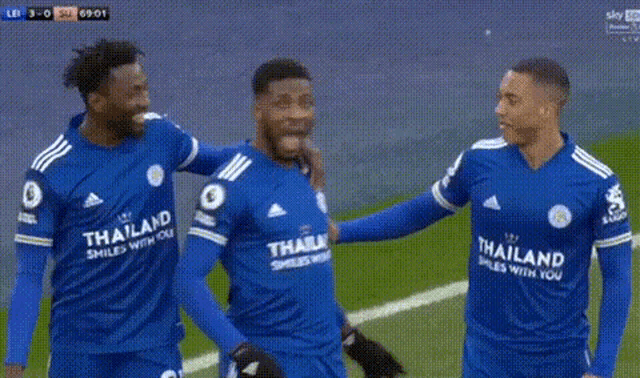 three soccer players wearing blue thailand smiles with you jerseys are standing on the field .