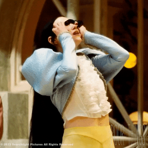 a woman wearing a blue jacket and yellow pants is standing in front of a sign that says poor things