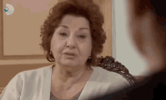 an older woman sitting in a chair talking to another woman with a tv channel hanging from the wall behind her