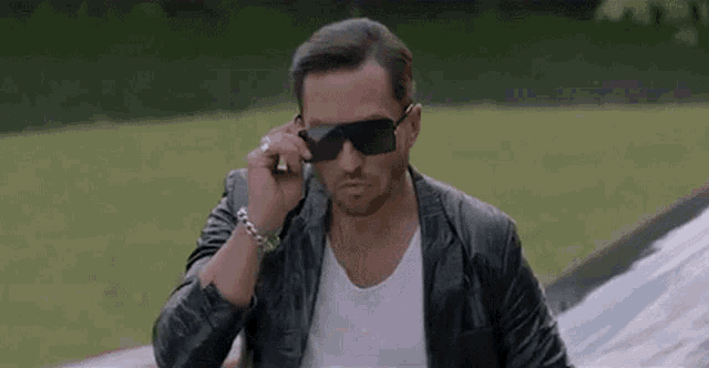 a man wearing sunglasses and a leather jacket is talking on a cell phone in front of a car .