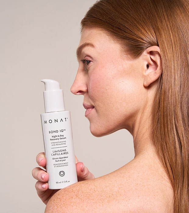 Profile view of a woman with straight, honey-blonde hair pulled back, holding a MONAT BOND IQ serum bottle