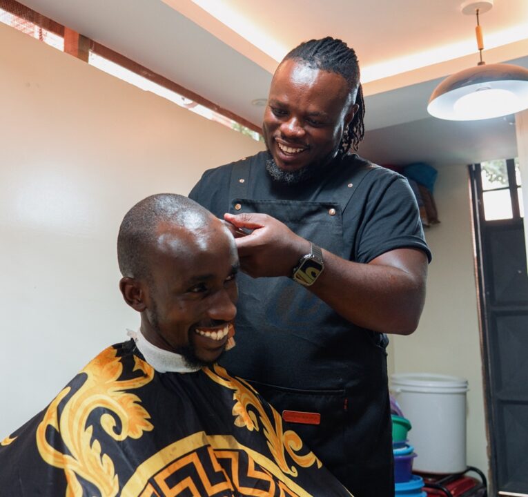 A man, dressed in black with a black apron, passes an electric razor over another man’s head.