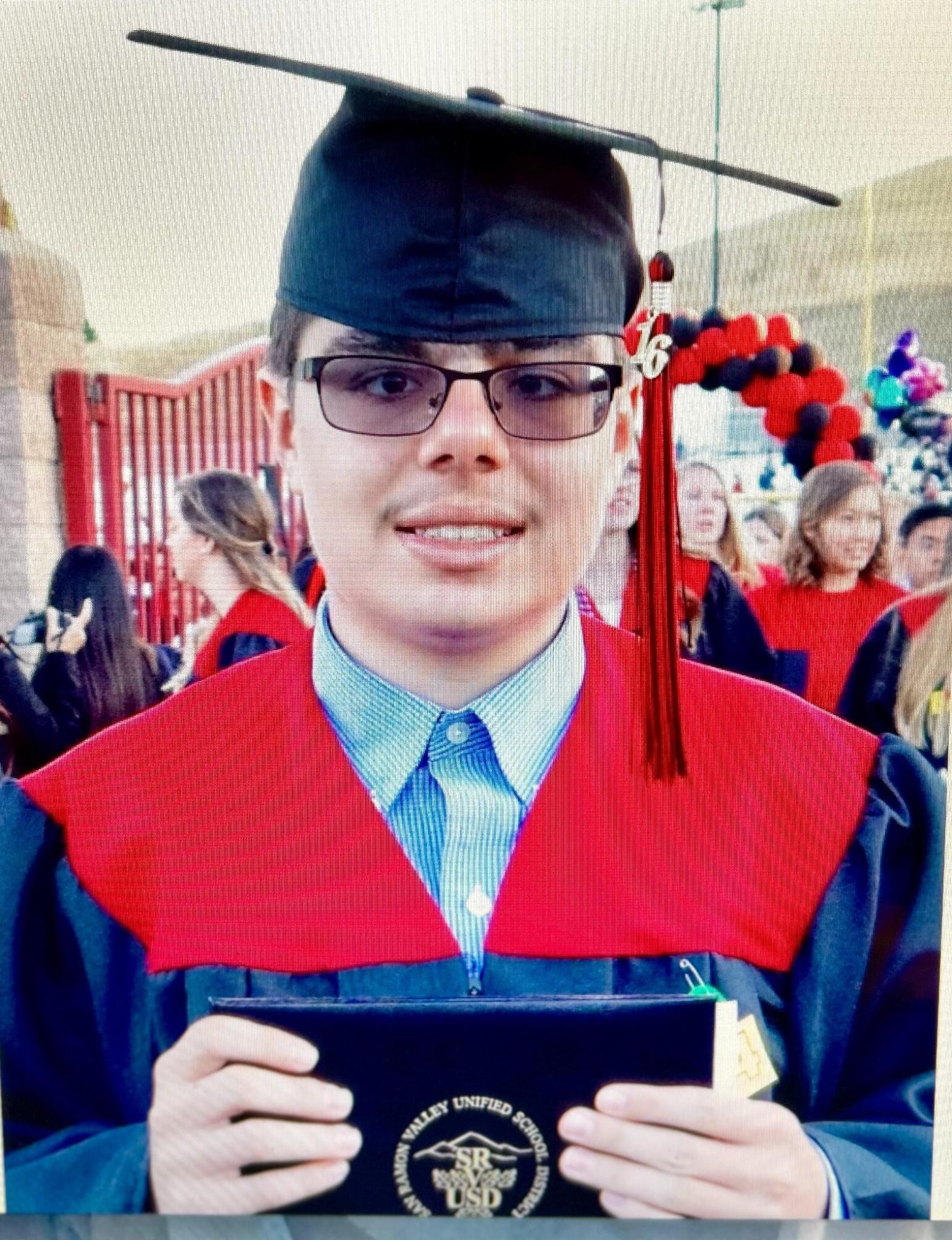 A young adult wearing glasses and a black graduation cap with a tassel reading “16” stands in front of a crowd at a commencement ceremony. He’s dressed in a robe with a red stole, over a collared button-down shirt, and holds a black diploma cover bearing the seal of the San Ramon Valley Unified School District. A red and black balloon arch and other graduates are visible in the background.