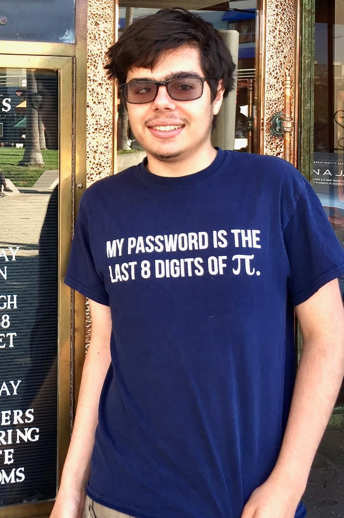 A young adult with short, dark hair and tinted glasses smiles at the camera. He wears a navy blue T-shirt with white text that reads, “MY PASSWORD IS THE LAST 8 DIGITS OF π.” He is standing in front of a brass-framed entrance, with part of the building’s signage visible behind him.