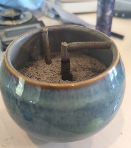 An incense censer with a burning Tibetan style stick held upright be incense ash. A segment of ash has fallen to form a bridge between the incense and the side wall of the censer.