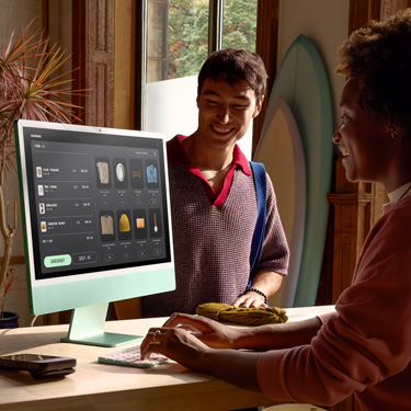Egy szörfdeszkákat és más termékeket árusító bolt alkalmazottja a pult mögött ül, és iMacen véglegesíti a pult másik oldalán álló vásárló tranzakcióját.
