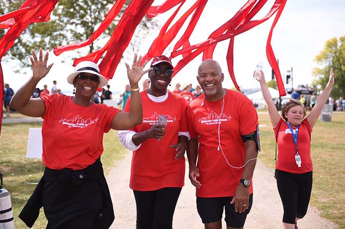 A group of smiling people wearing red is walking down a sidewalk and cheering.