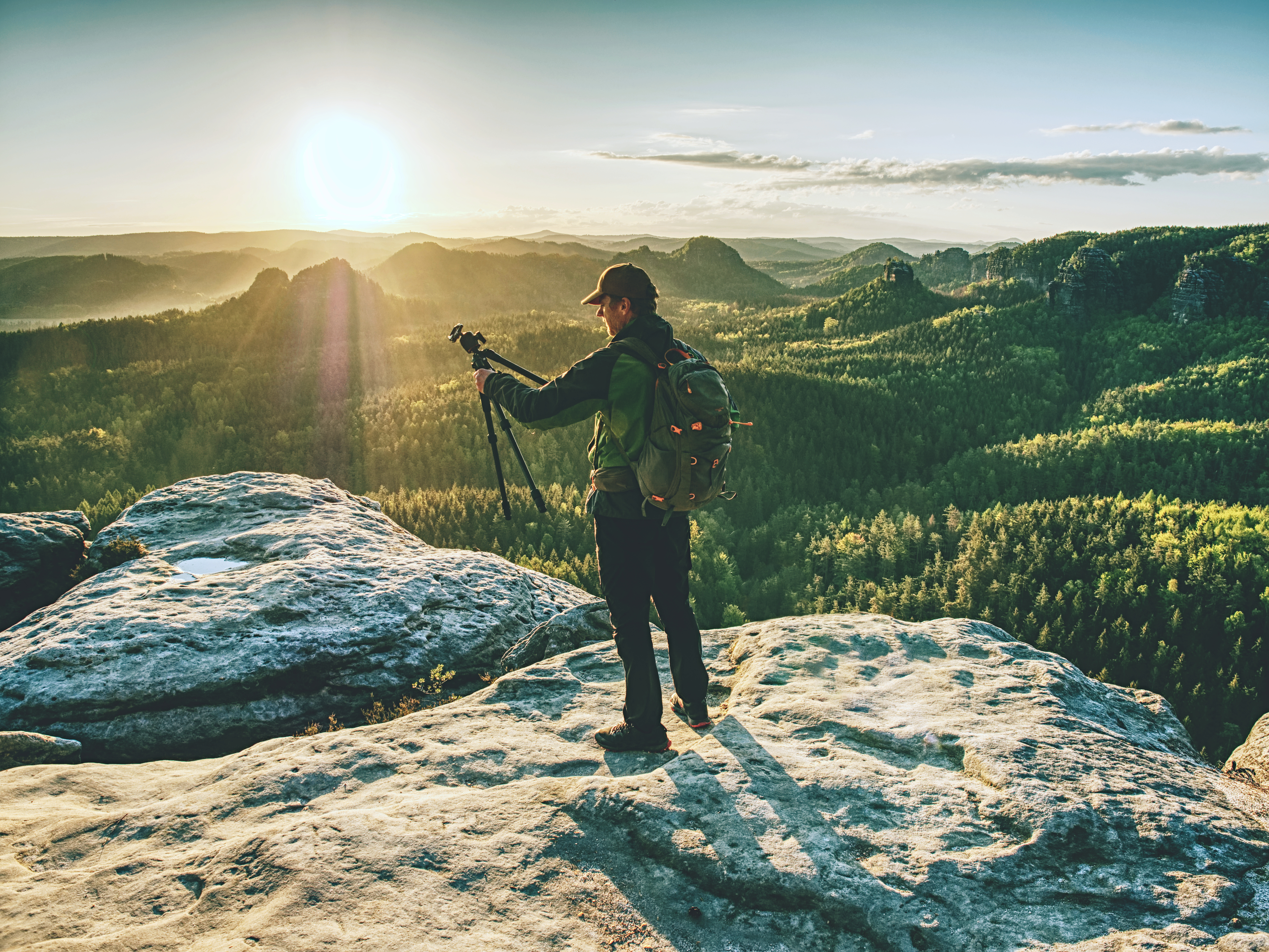 man in outdoor style clothing holding a tripod and stood atop a mountain
